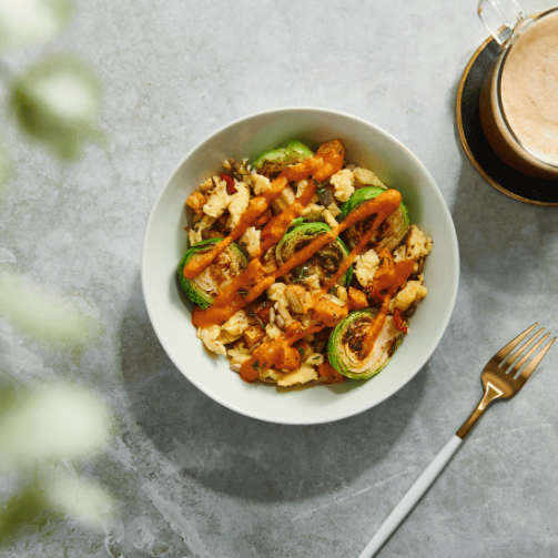 Eine weiße Schale mit einem Gericht aus dem Modern-Recipe-Foodkonzept: Rosenkohl mit Gemüse und Sauce. Daneben eine Gabel und eine Tasse Kaffee.