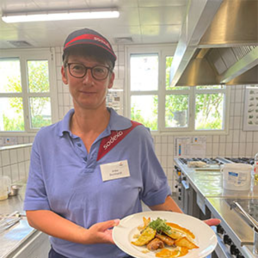 Portrait von Anke Bernhardt, Köchin bei Sodexo, die in einer Großküche ein Gericht präsentiert