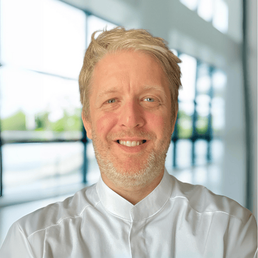 Portrait von Reuven Schockner, Head of Operational and Culinary Excellence bei Sodexo Deutschland in Kochjacke vor einer Fensterfront.