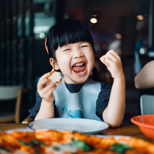kleines asiatisches Mädchen, das glücklich in die Kamera lacht und ein Stück Pizza in der Hand hält.