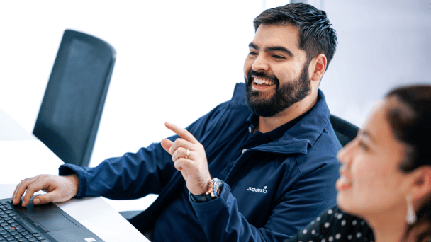 Ein Mitarbeiter von Sodexo sitzt an einem Konferenztisch, lacht und hat seinen Finger erhoben. Im Vordergrund sieht man im Anschnitt eine Frau sitzen.