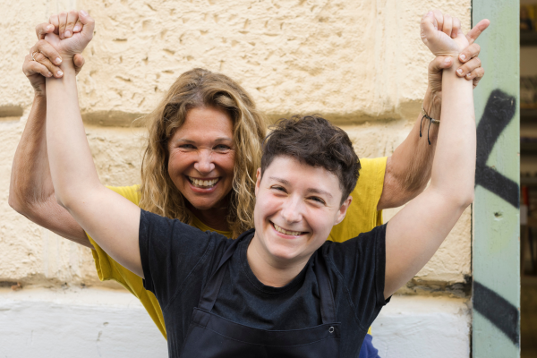 Haya Molcho steht hinter Sodexo-Köchin Fausta Tortoriello. Beide lachen in die Kamera und heben ihre Arme in Siegerpose nach oben.