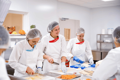 Sechs weibliche Sodexo-Küchenmitarbeiterinnen mit Sodexo-Schürzen, Haarnetzen und Handschuhen, die Gemüse schneiden und vorbereiten.