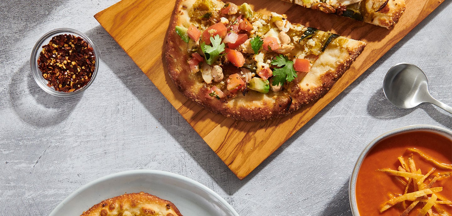 Ein lecker belegtes Stück Pinsa serviert auf einem Holzbrett. Daneben eine Schüssel Chiliflocken und eine Tomatensuppe