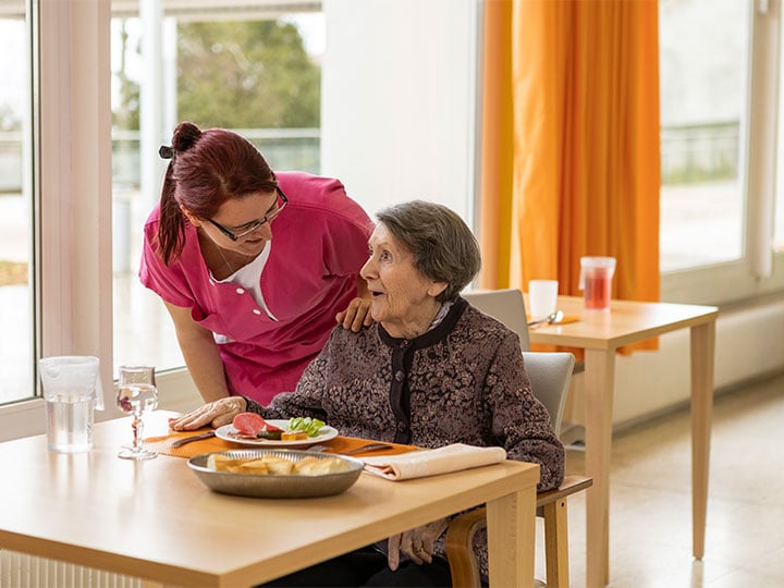 Mitarbeiterin einer Senioreneinrichtung kümmert sich freundlich um Bewohnerin, die lachend am Tisch, gedeckt mit Essen, sitzt