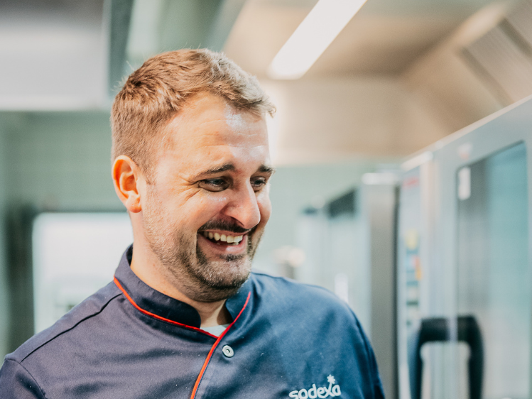 Sodexo Food-Experte Jascha Händle in Nahaufnahme lachend in Kochuniform in einer Großküche.