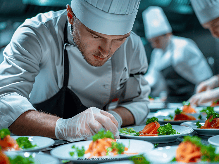 Ein Koch in Kochjacke und Mütze dekoriert eine Reihe an Salattellern mit Kräutern.