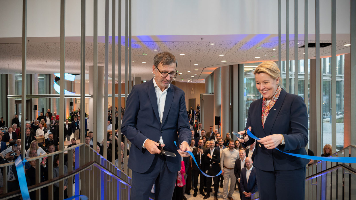 Franziska Giffey und ein Mann im Anzug durchschneiden gemeinsam ein blaues Band bei der Gebäudeeröffnung, dahinter zahlreiche Gäste im Foyer.