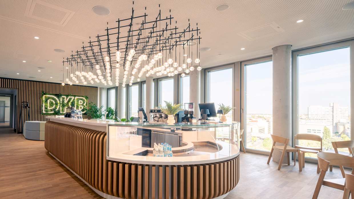 Geschwungene Kaffeebar aus Holzlamellen mit gläserner Auslage, darüber eine große Leuchteninstallation. Im Hintergrund ein begrüntes DKB-Logo an einer Lamellenwand.