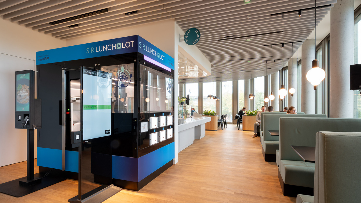 Küchenroboter Sir Lunchalot von goodBytz mit blauem Display und Abholfächern im Betriebsrestaurant, rechts grüne Sitznischen.