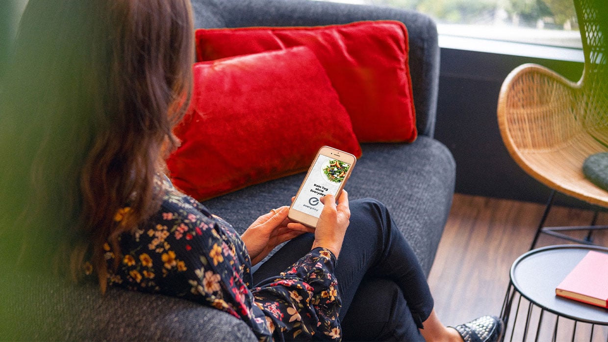 Blick über die Schulter einer Frau, die in ihren Handy ein Smartphone mit der geöffneten Everyday by Sodexo-App hält.