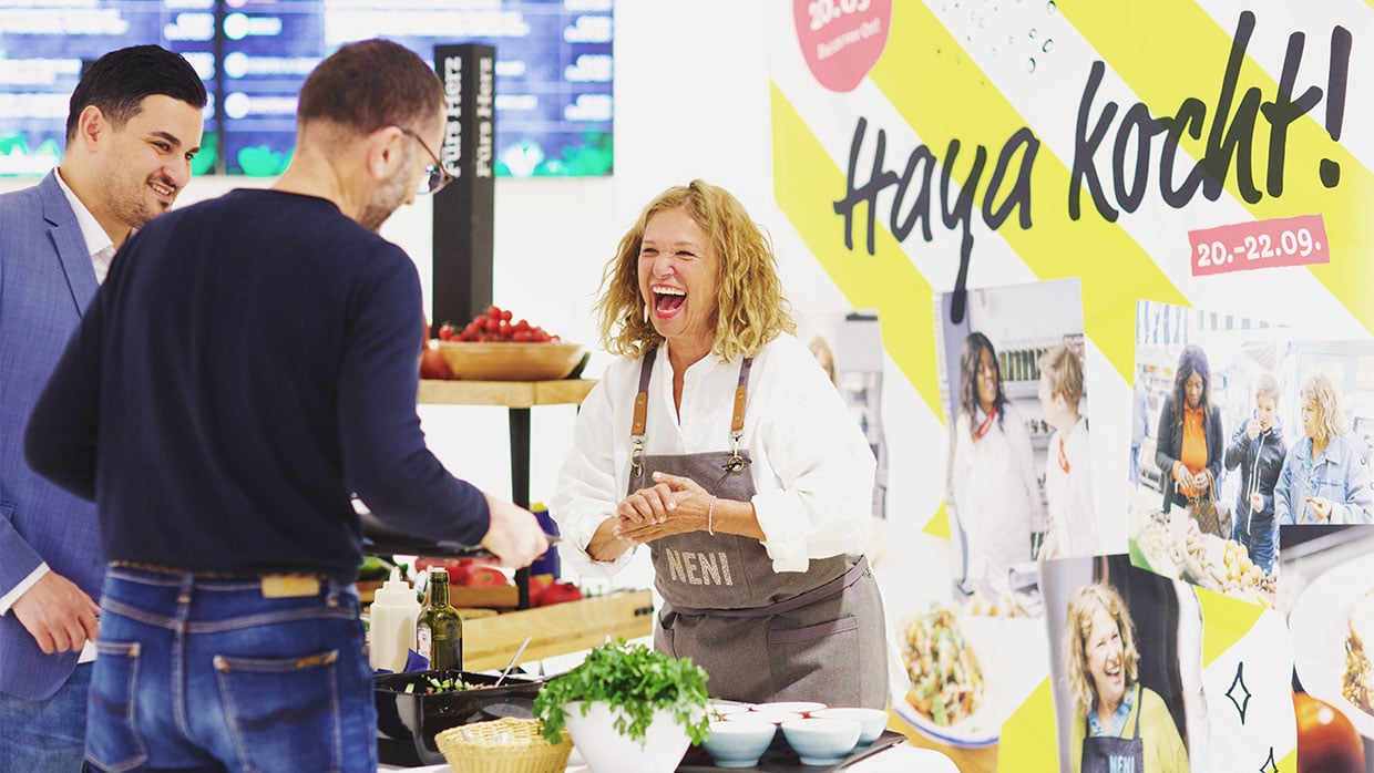 Haya Molcho (Neni) lacht beim Sodexo-Kochevent mit zwei männlichen Gästen bei der Essensausgabe