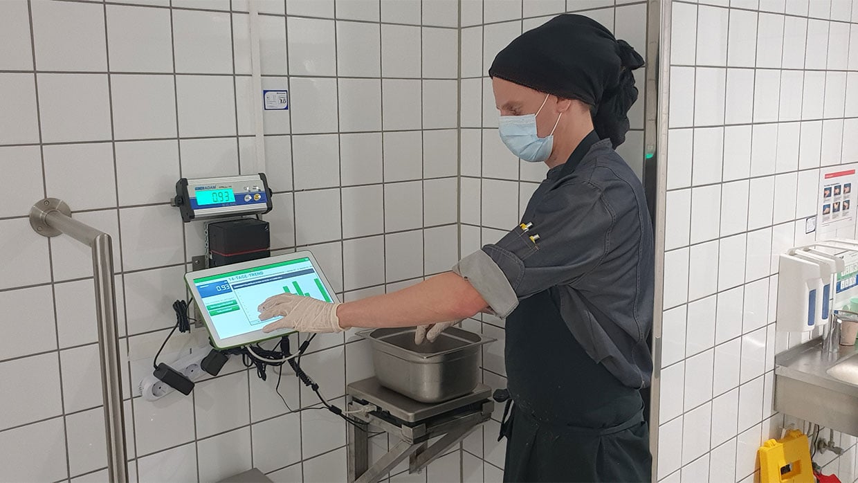 Sodexo-Mitarbeiter mit Mundschutz bedient WasteWatch-Messgerät in einem Betriebsrestaurant, um Lebensmittelabfälle zu wiegen.