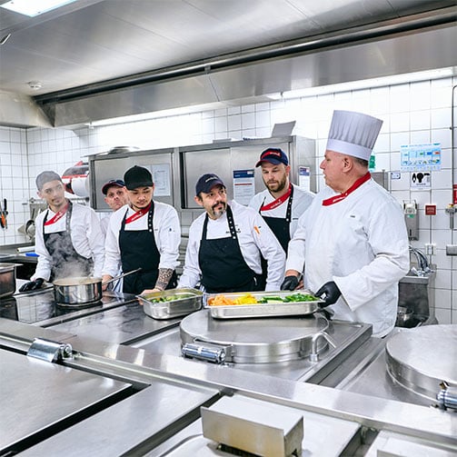 Sodexo-Betriebsleiter Thomas Klatt im Gespräch mit seinem Küchenteam in der Betriebsküche. In seiner Hand hält er einen Behälter mit Gemüse.