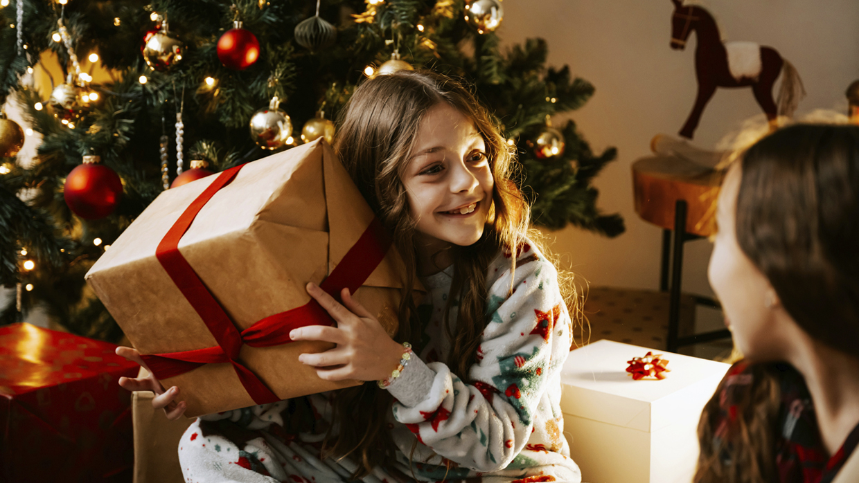 Hymyilevä lapsi joulukuusen ääressä lahjapaketin kanssa.