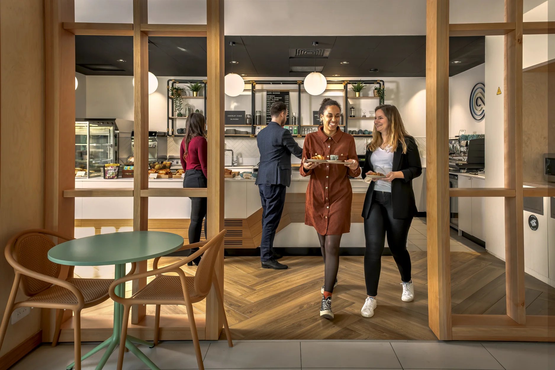 Generating responseCopilot said: Alt text: Employees collect refreshments and walk through a modern workplace café with wooden flooring, pendant lighting and a central coffee bar. Tables, chairs, display cabinets and coffee equipment are visible, creating a welcoming break-out space for dining and socialising.
