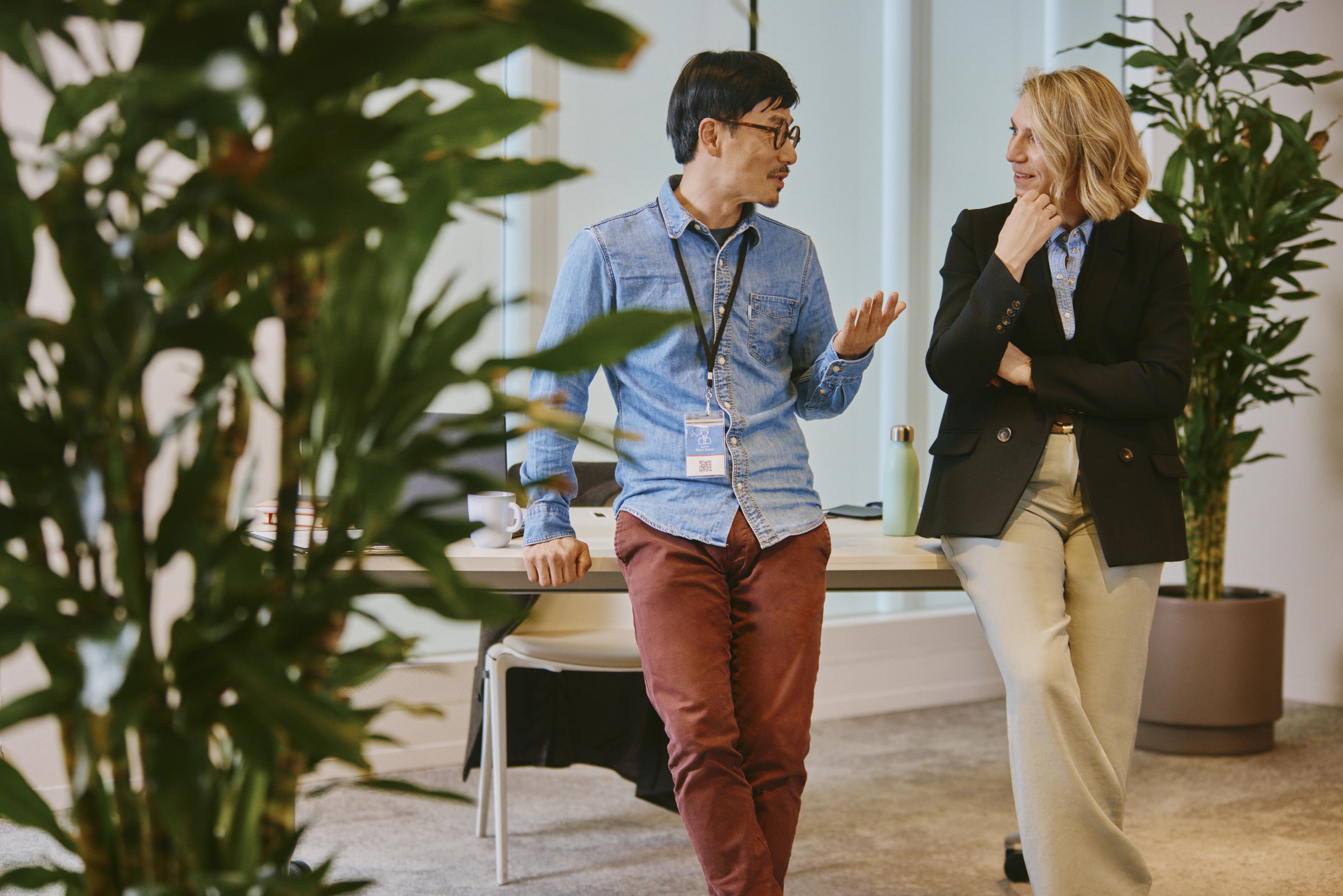 Two office workers walking through a bright workspace surrounded by large indoor plants. One person wears a blue shirt with a lanyard and red trousers, while another wears a dark blazer and light-coloured trousers. Desks and office furnishings are visible in the background.