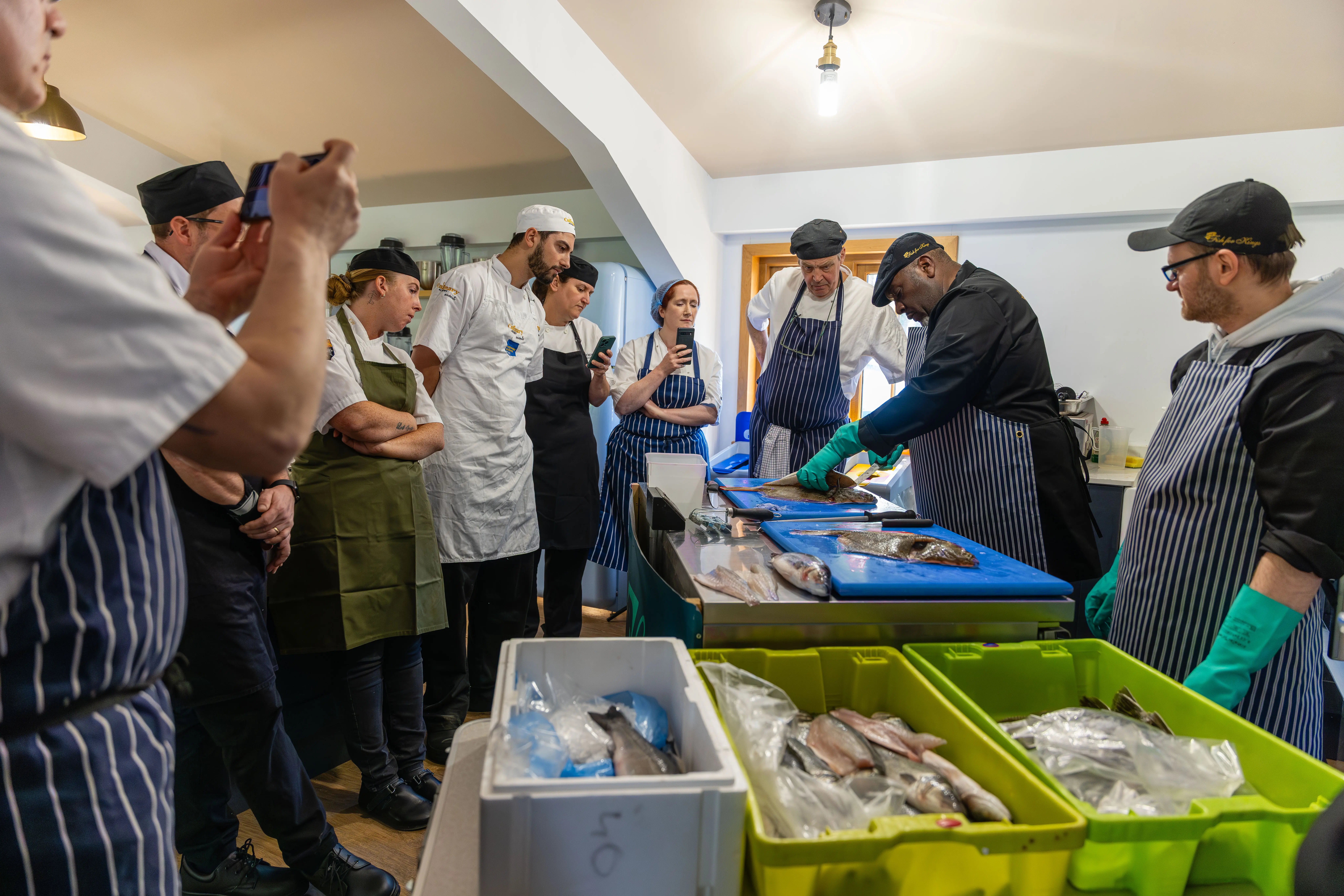 Indoor cooking workshop scene showing a group of people gathered around a long table in a kitchen. In the foreground, a person stands at the table preparing ingredients on a blue cutting board, surrounded by green bins filled with produce and containers of seafood. Other participants line the sides of the table wearing aprons and observing the demonstration, while overhead lights and kitchen equipment are visible in the background.
