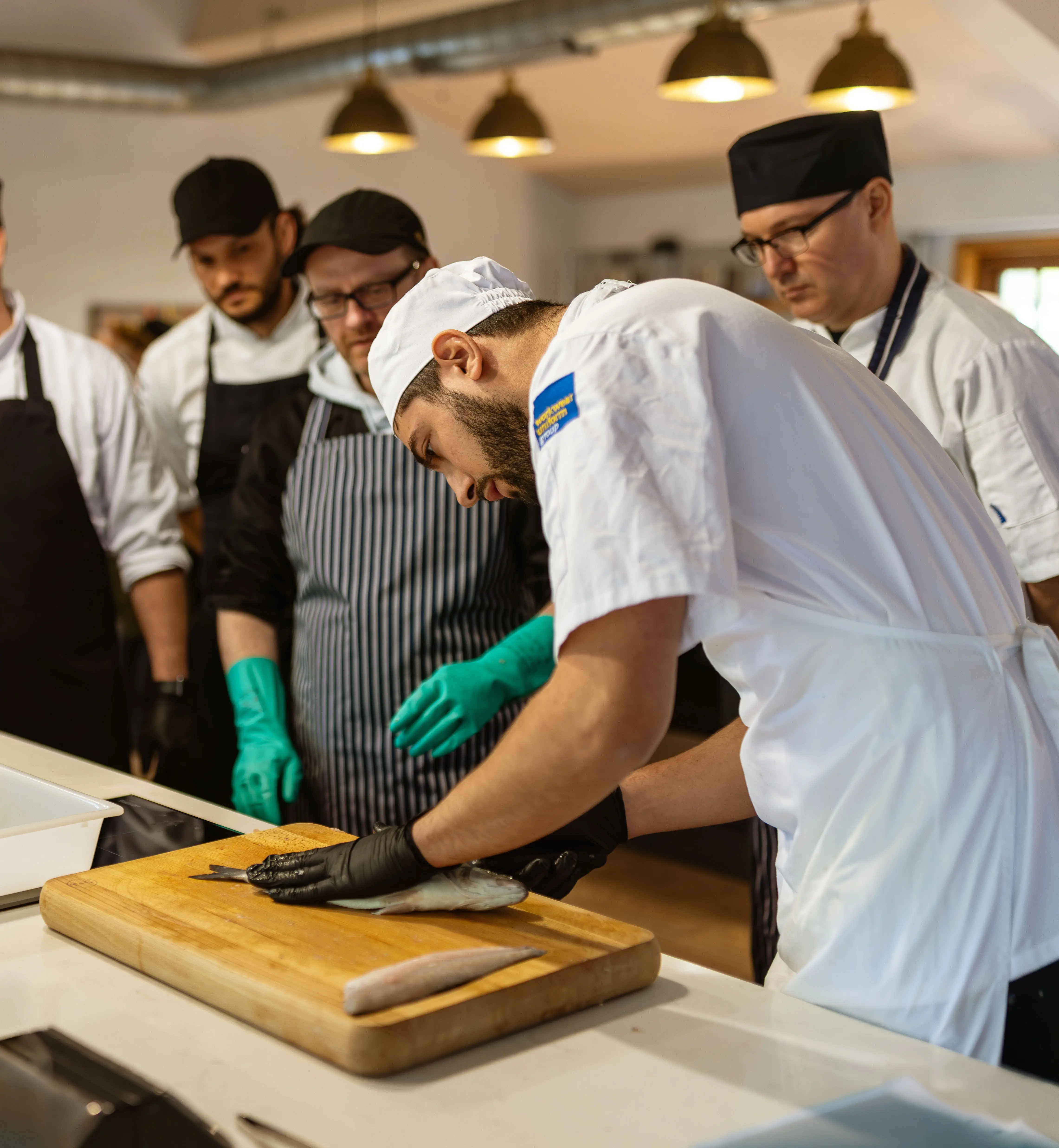 Commercial kitchen scene showing several people in chef uniforms and aprons gathered around a preparation area. In the foreground, one person wearing a white shirt, apron, cap, and blue gloves is working at a wooden cutting board with utensils and containers nearby, while others stand behind observing and assisting.