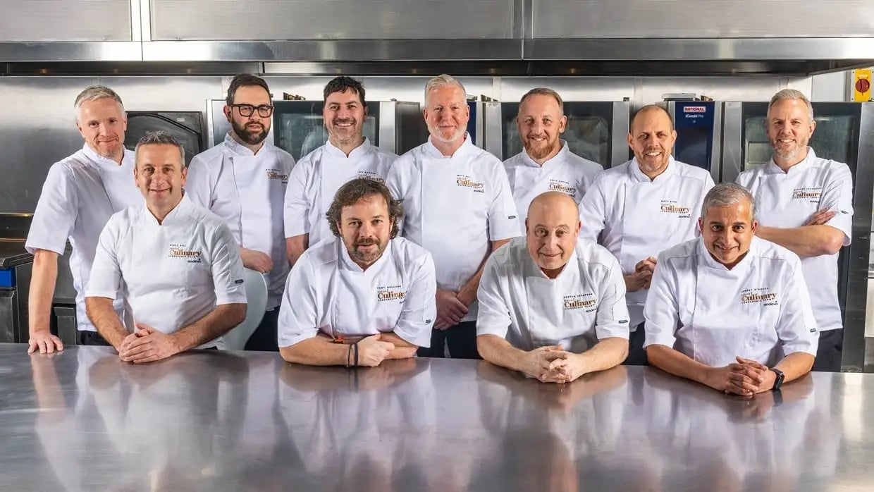 A group of chefs in white uniforms standing and leaning around a stainless steel kitchen counter in a professional kitchen setting