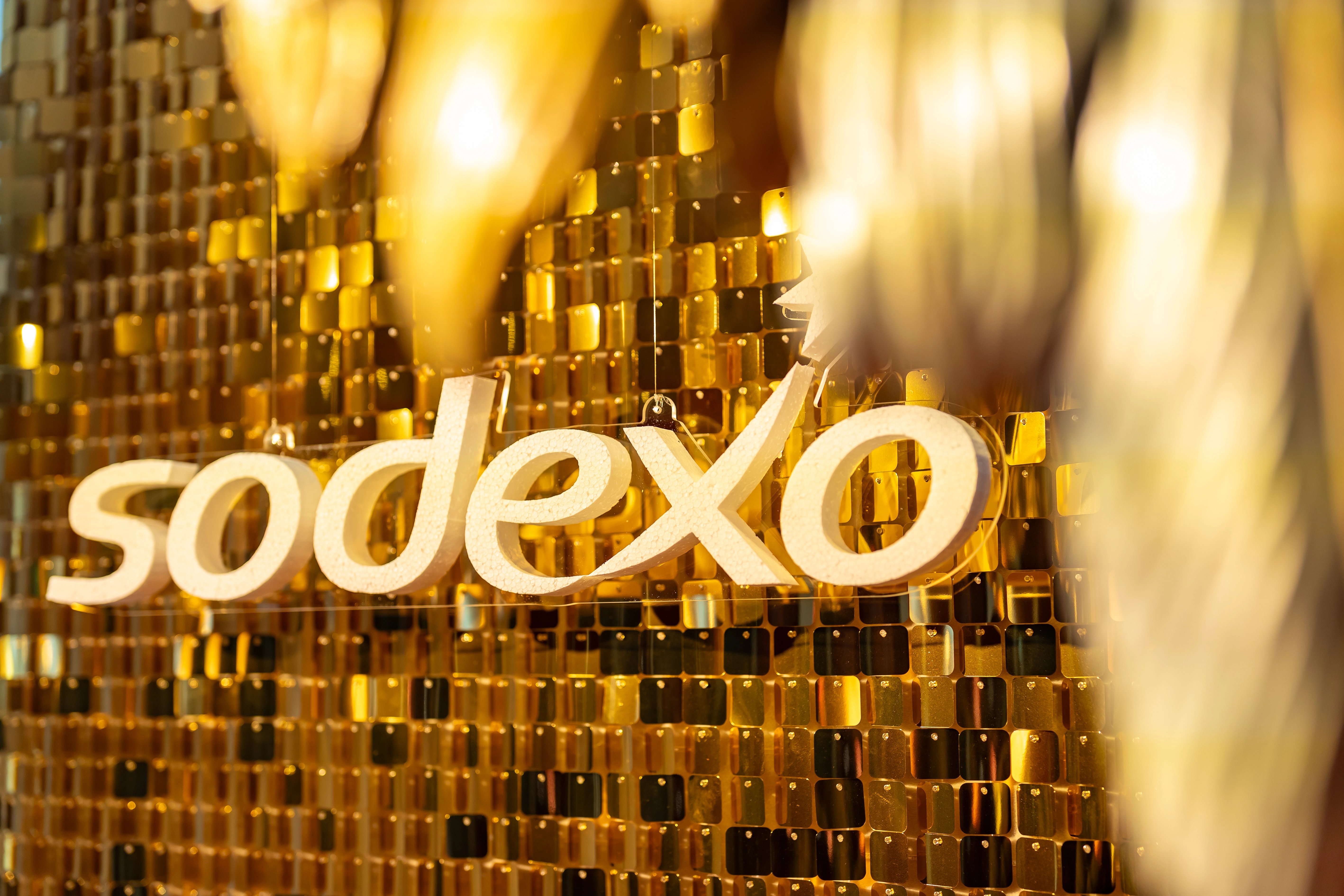 Sodexo wordmark displayed against a gold mosaic background with warm, defocused lighting creating a festive, upscale atmosphere.