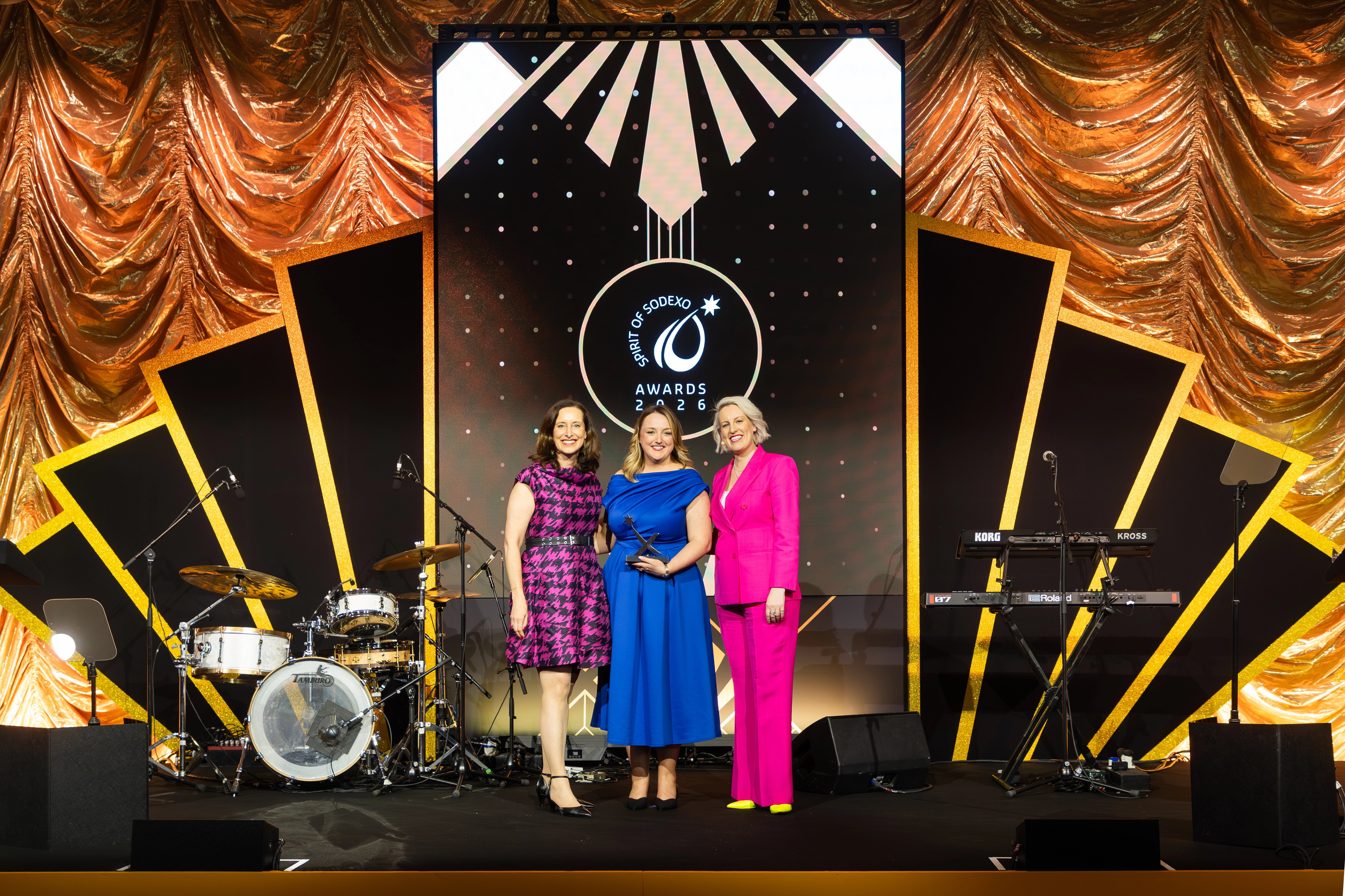 Three attendees stand on a stage at the Spirit of Sodexo Awards 2026, with one holding a trophy, framed by gold draped curtains, musical instruments, and a large branded screen backdrop.