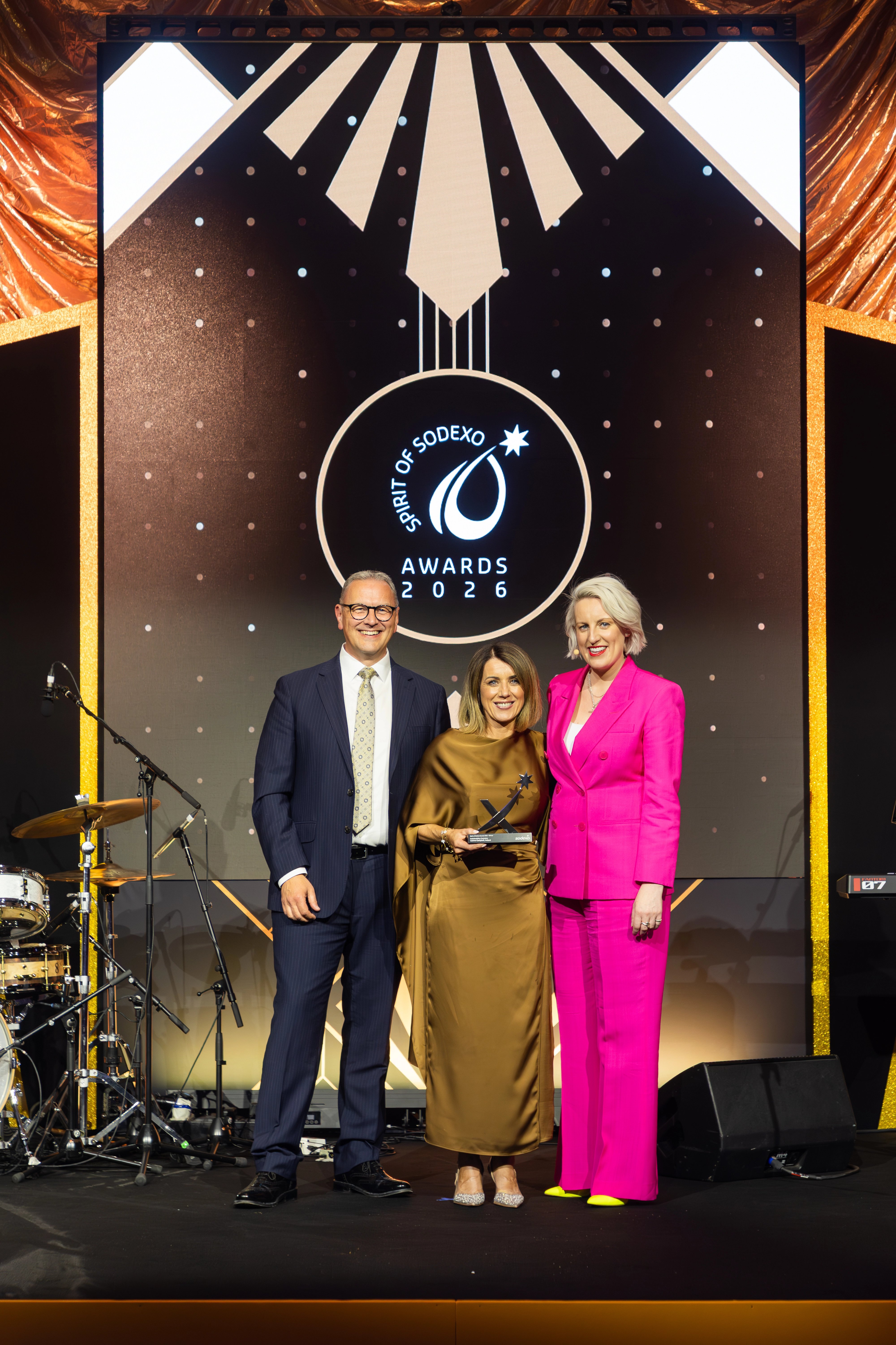 Three attendees stand on stage at the Spirit of Sodexo Awards 2026, with one holding a trophy, in front of a black and gold art deco–style branded screen and gold draped backdrop.