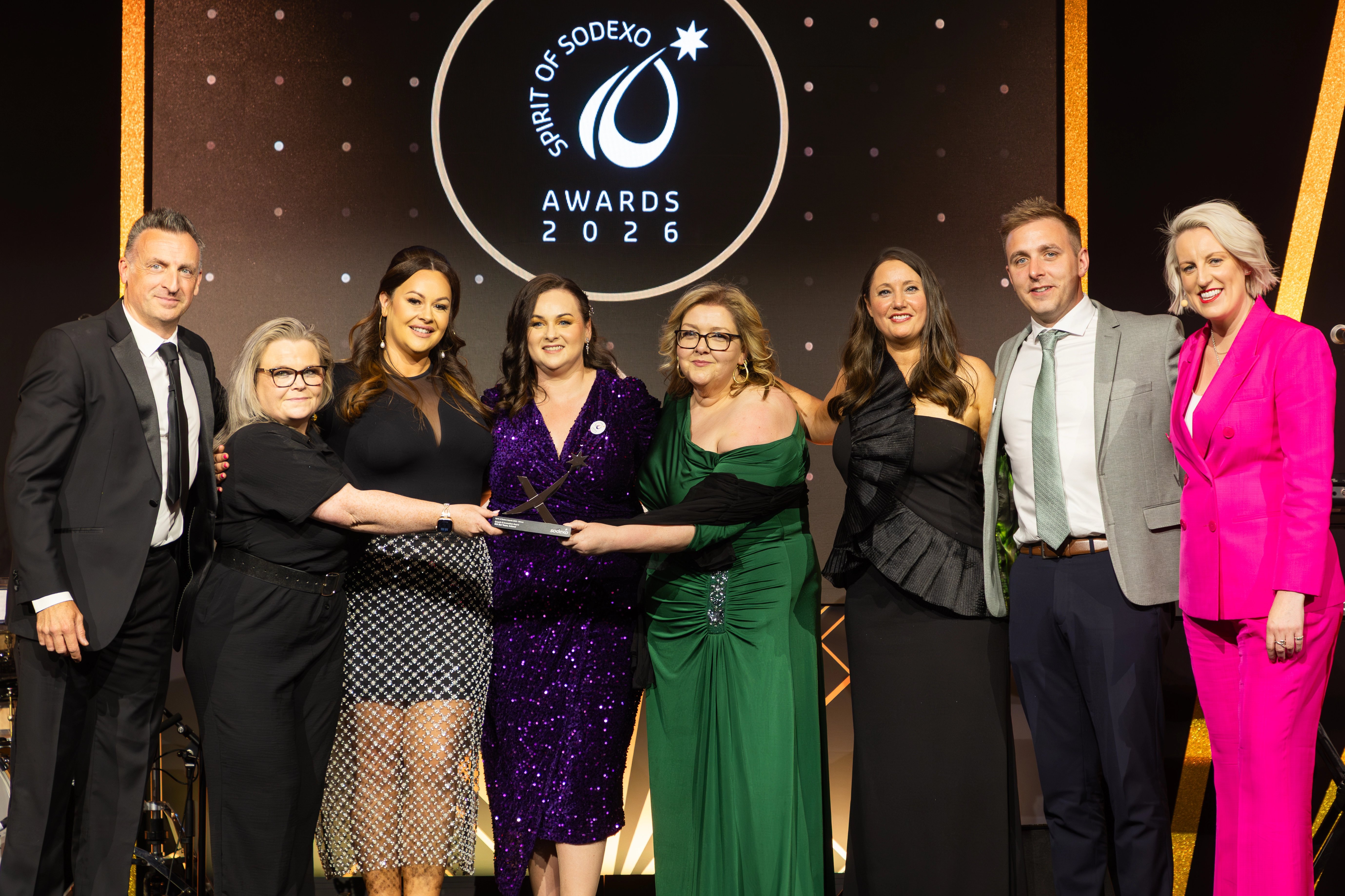 Group of attendees on stage at the Spirit of Sodexo Awards 2026 holding an award trophy together, dressed in formal evening wear in front of a black and gold branded backdrop.