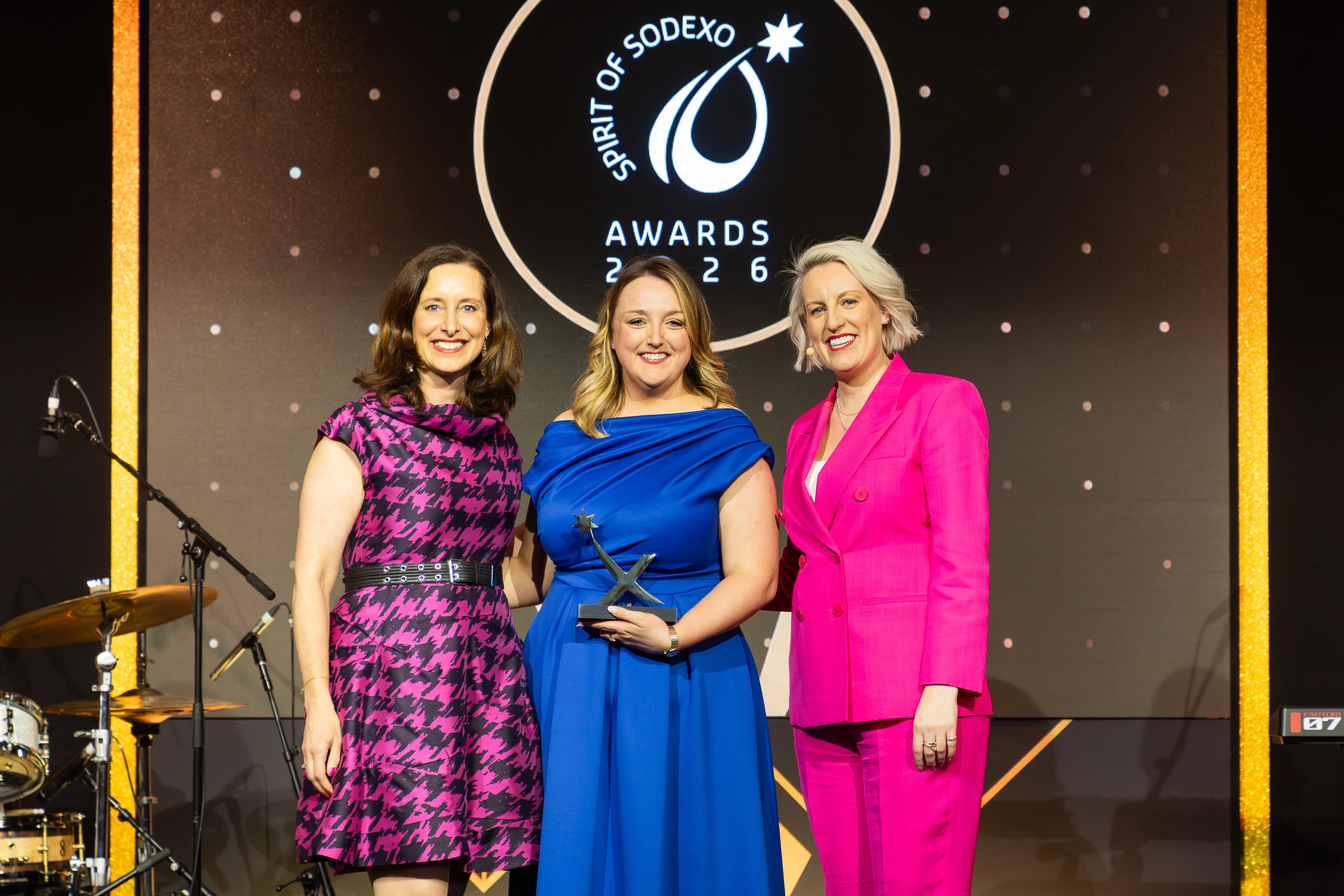 Three attendees stand on stage at the Spirit of Sodexo Awards 2026, posing with a trophy in front of a black and gold branded backdrop with event logo and spotlit staging.