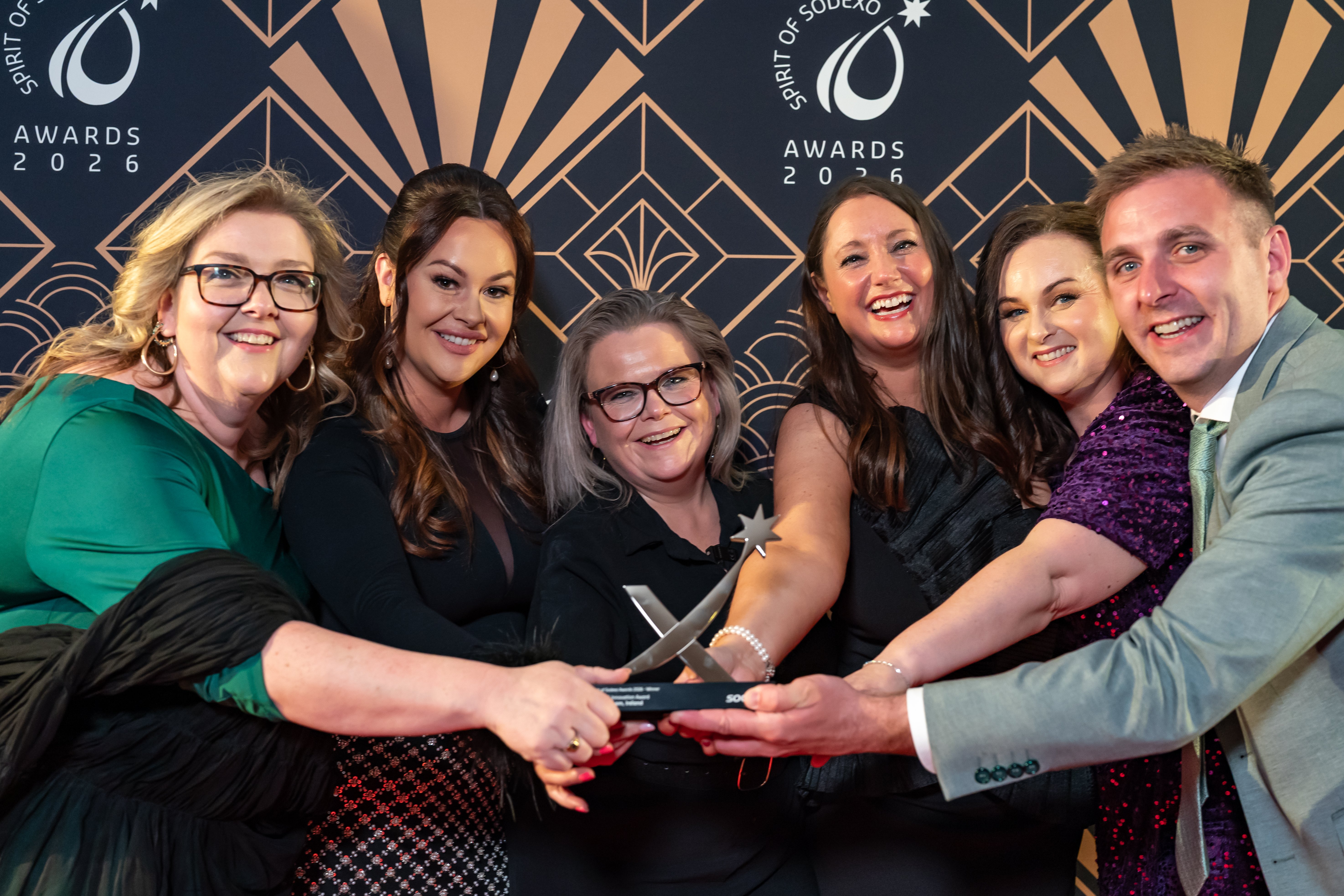 Group of six formally dressed attendees gathered around an award trophy, placing hands together in the centre, in front of a black and gold Spirit of Sodexo Awards backdrop with art deco
