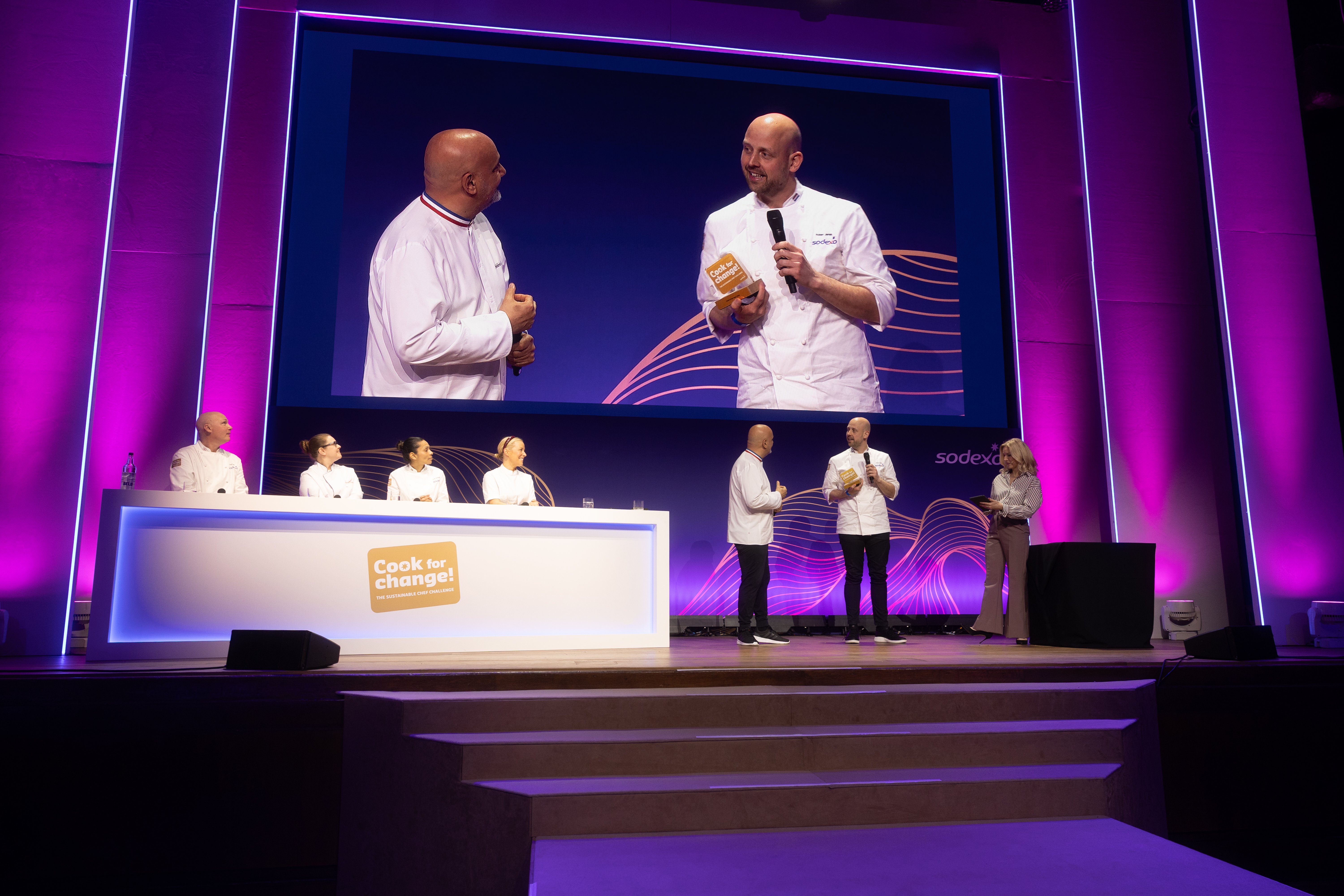 hefs stand on stage as one receives an award, with the scene shown on a large screen above. Other chefs sit at a judging table under “Cook for Change!” branding and purple lighting