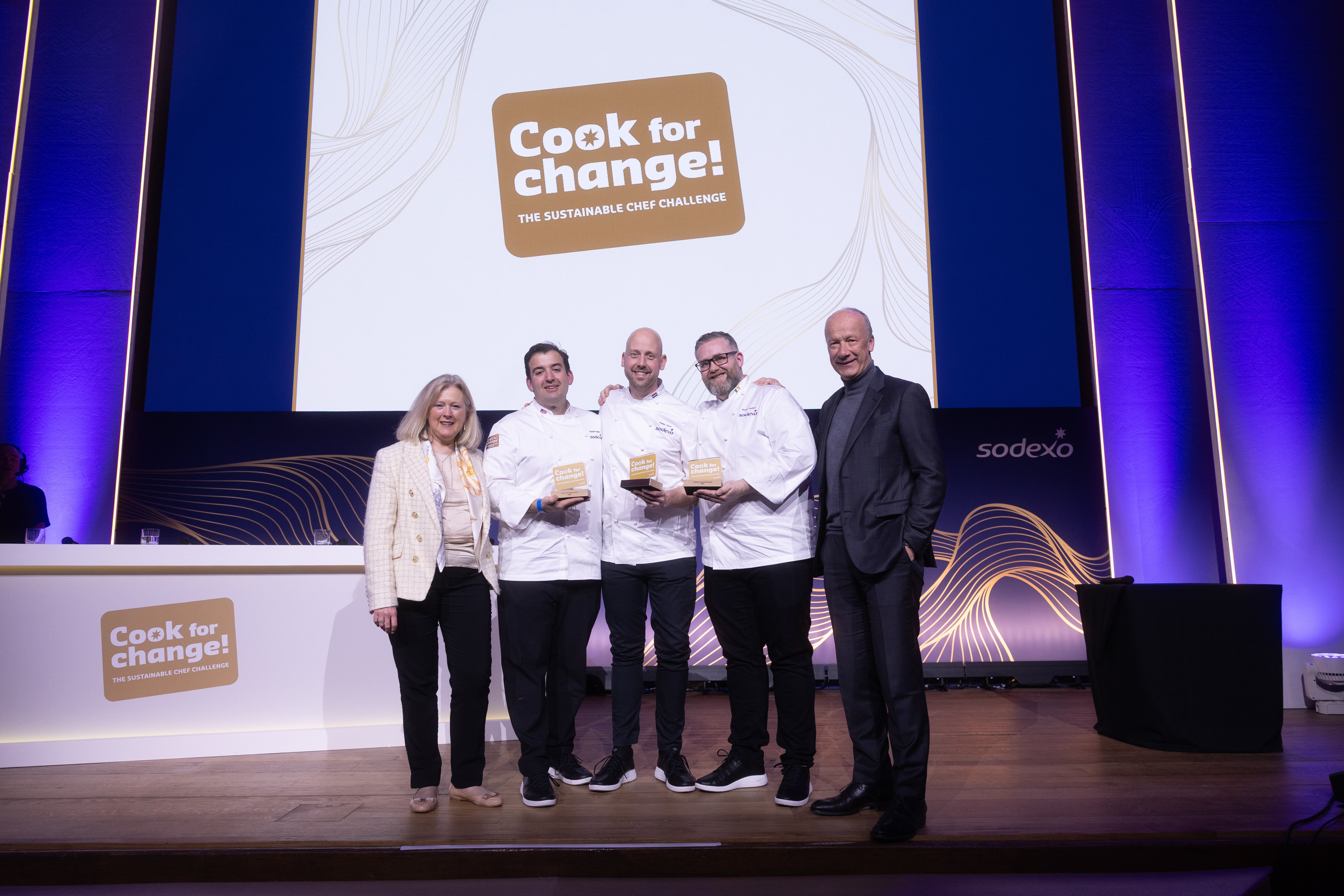 Three chefs holding awards stand on stage with two presenters, in front of a large “Cook for Change!” event screen.