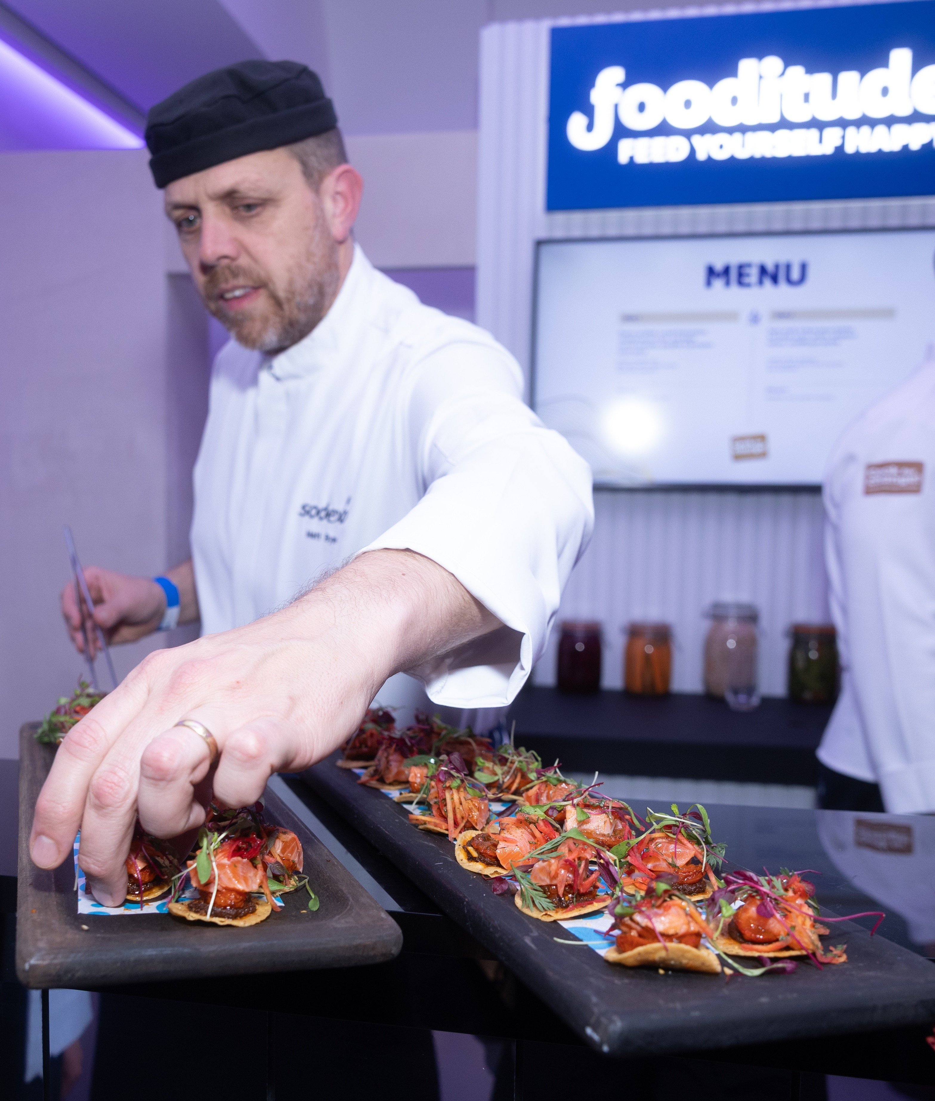 A chef in a white uniform arranges small plated appetizers at a food station with a “fooditude” menu sign in the background.