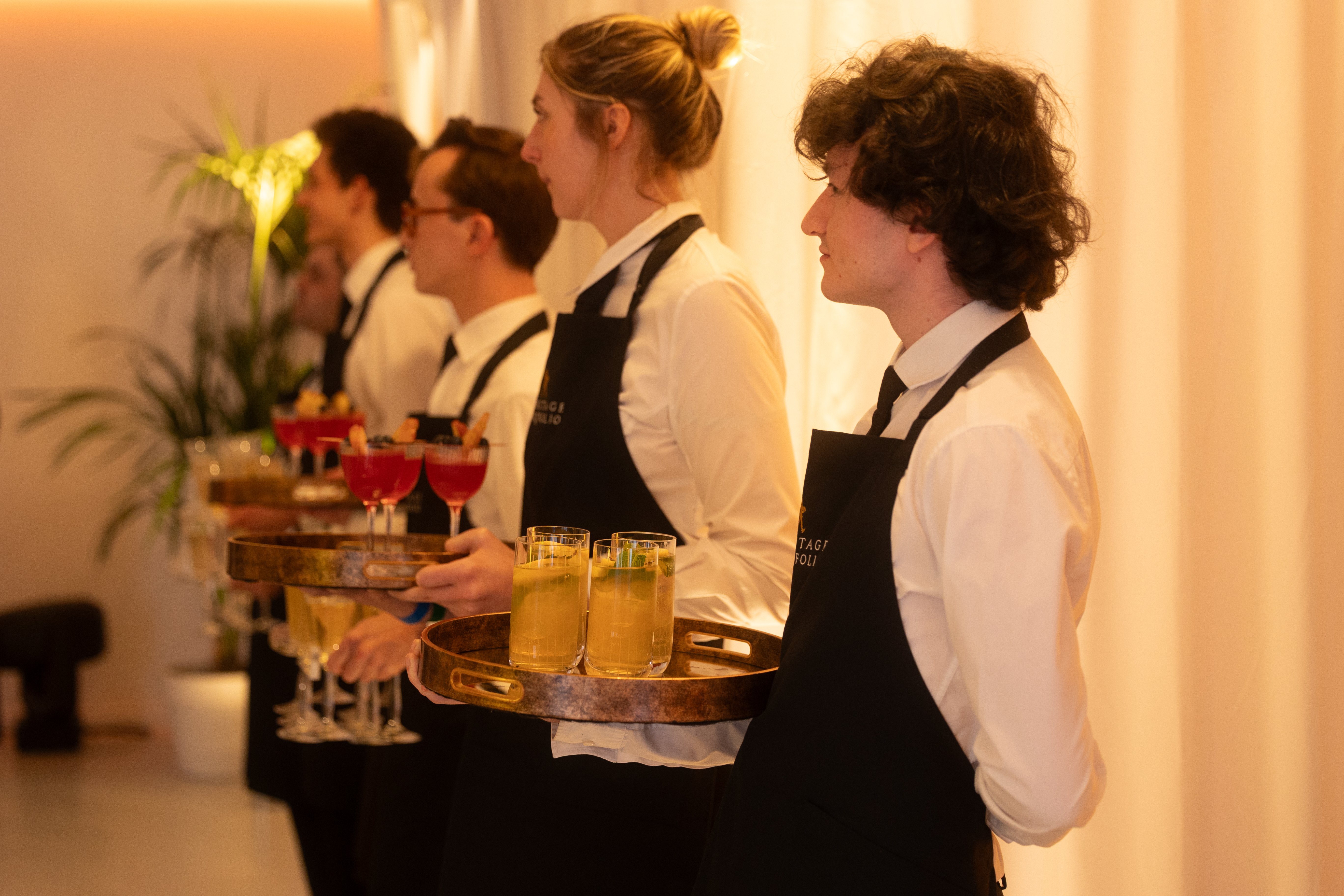 Servers in white shirts and black aprons stand in a line holding trays of assorted cocktails.