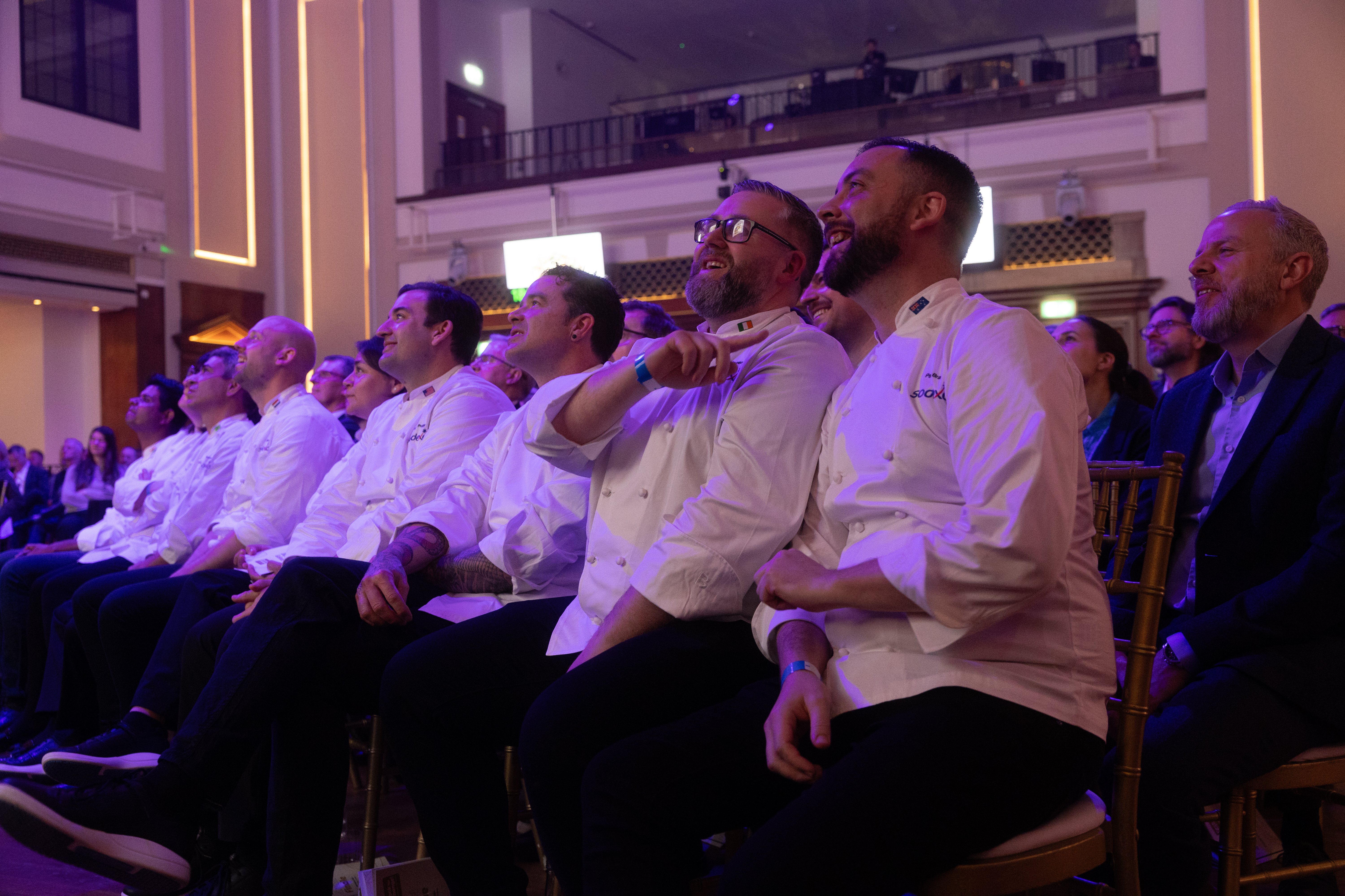 Chefs seated in an audience laugh and watch the stage during the Cook for Change event