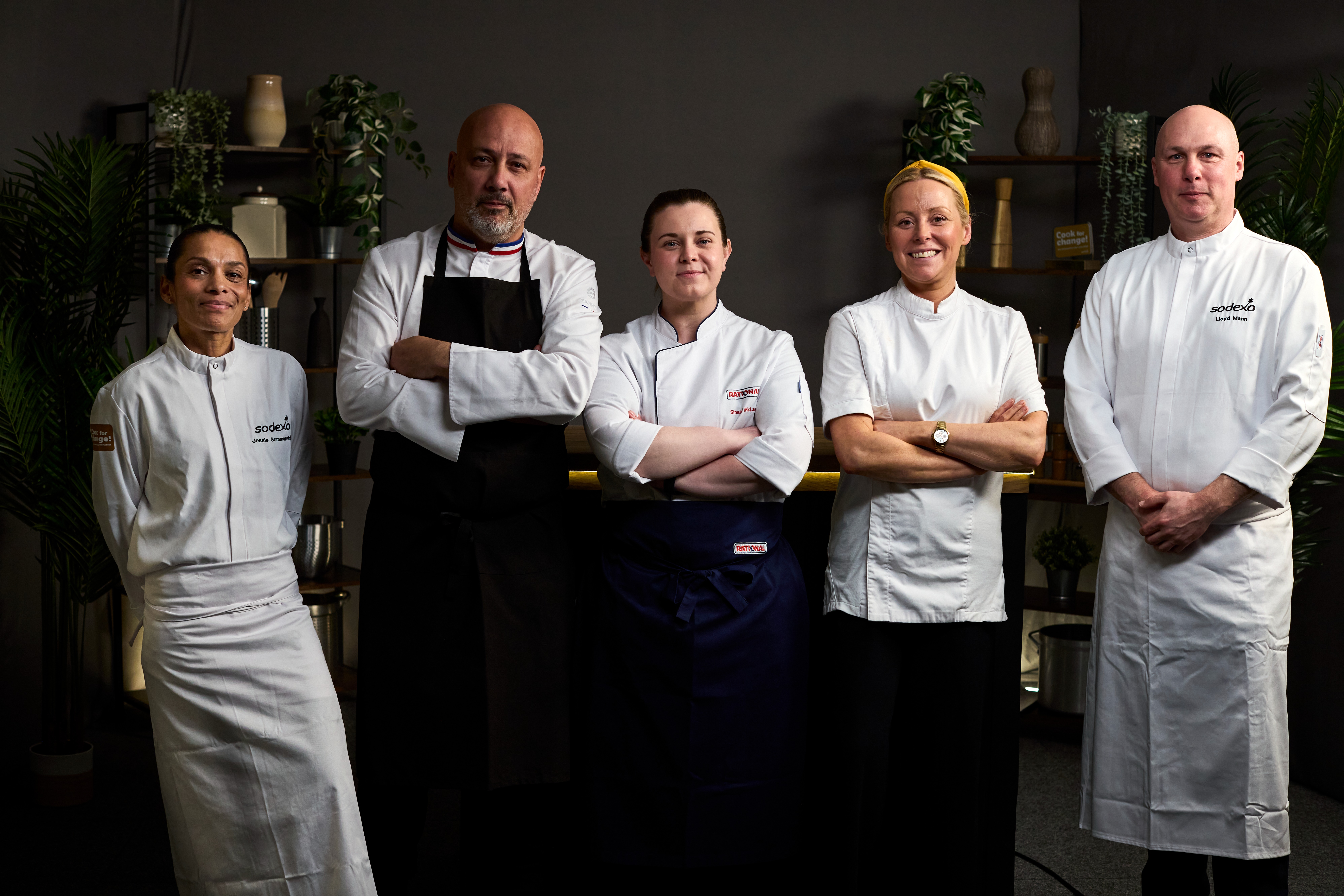 Five chefs stand side by side in a studio setting, posing confidently in their uniforms.