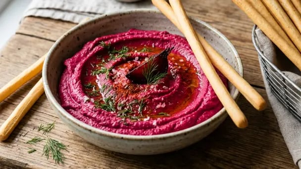 Roasted beetroot hummus in a speckled ceramic bowl, drizzled with olive oil and garnished with fresh dill and paprika, served with breadsticks on a wooden board