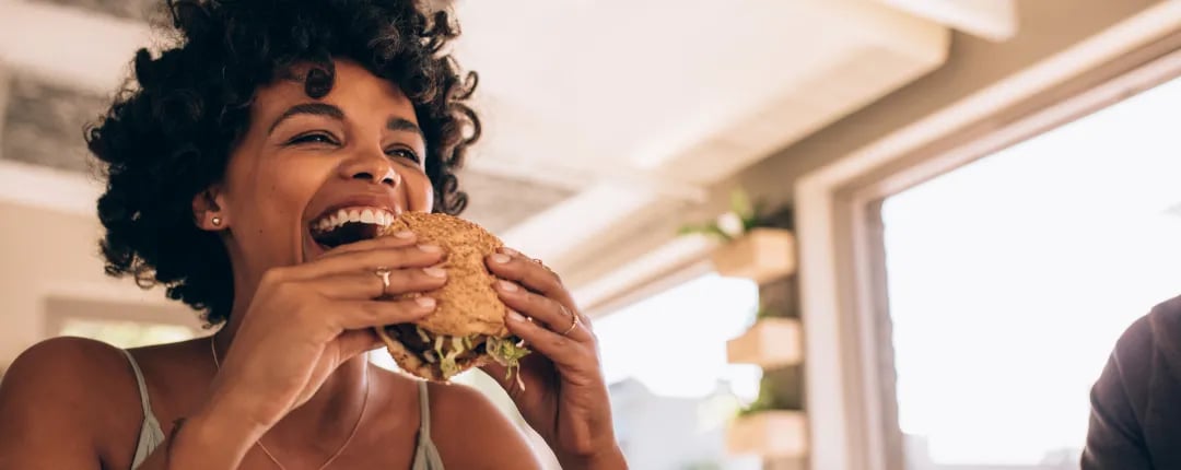 Person holding and eating a seeded sandwich indoors, with natural light streaming through large windows.