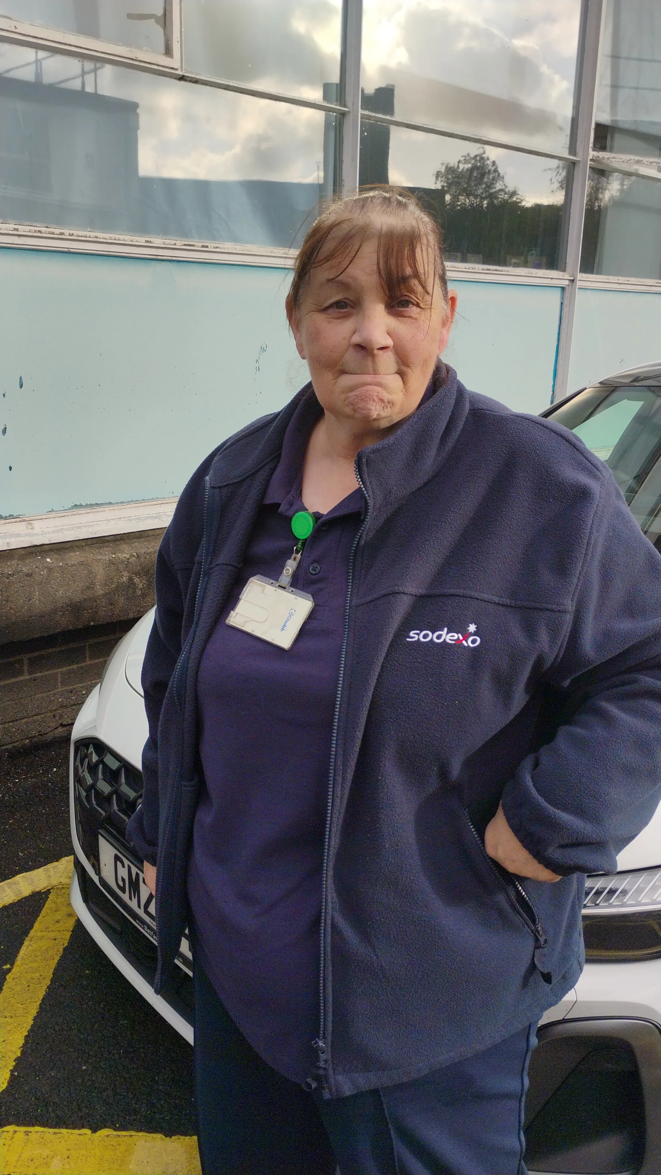 Sodexo colleague wearing a navy Sodexo fleece and ID badge, standing outdoors in front of a building and parked vehicle.