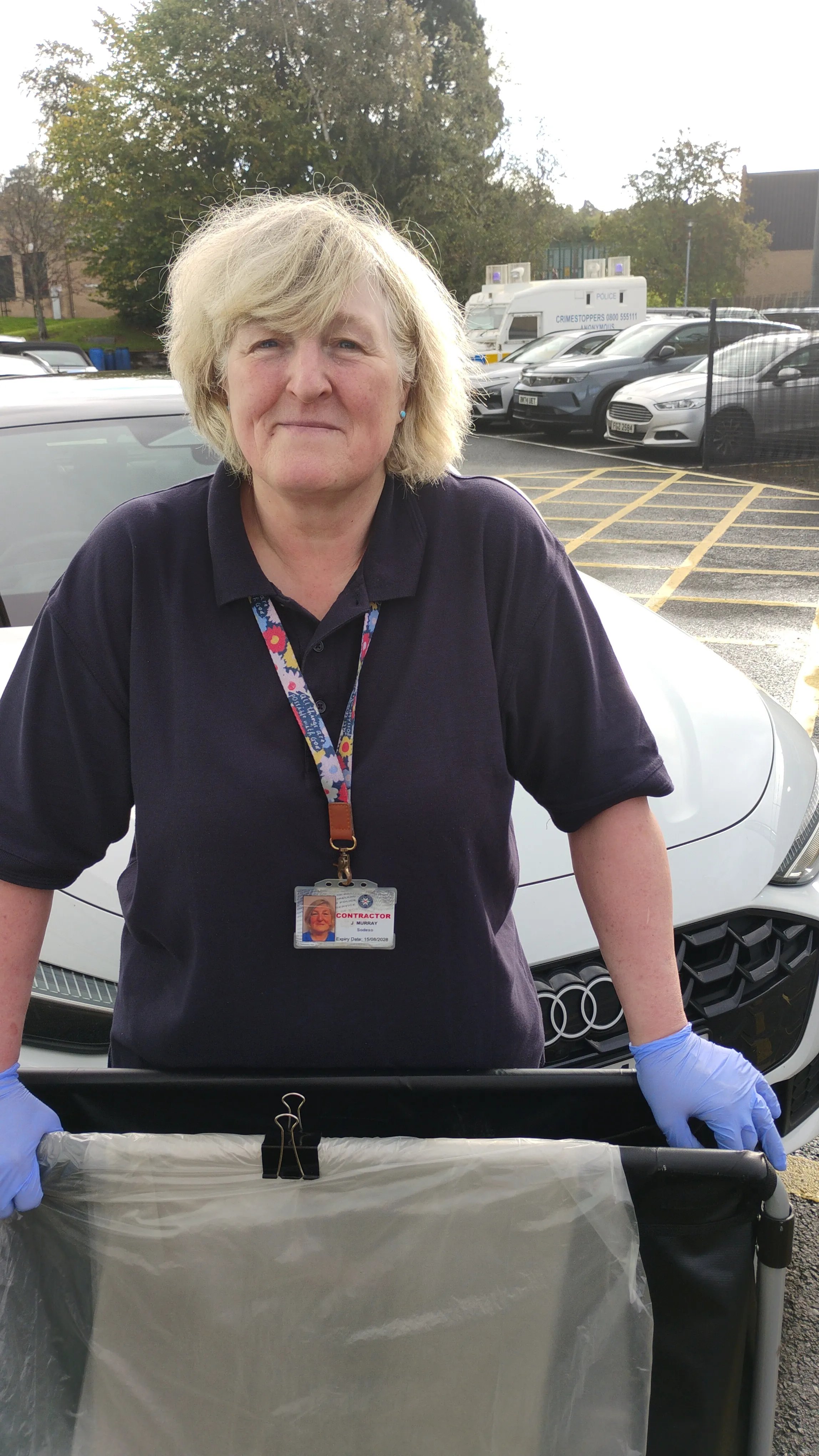 Colleague wearing protective gloves standing outdoors with a waste collection trolley in a car park, with vehicles and workplace buildings in the background.