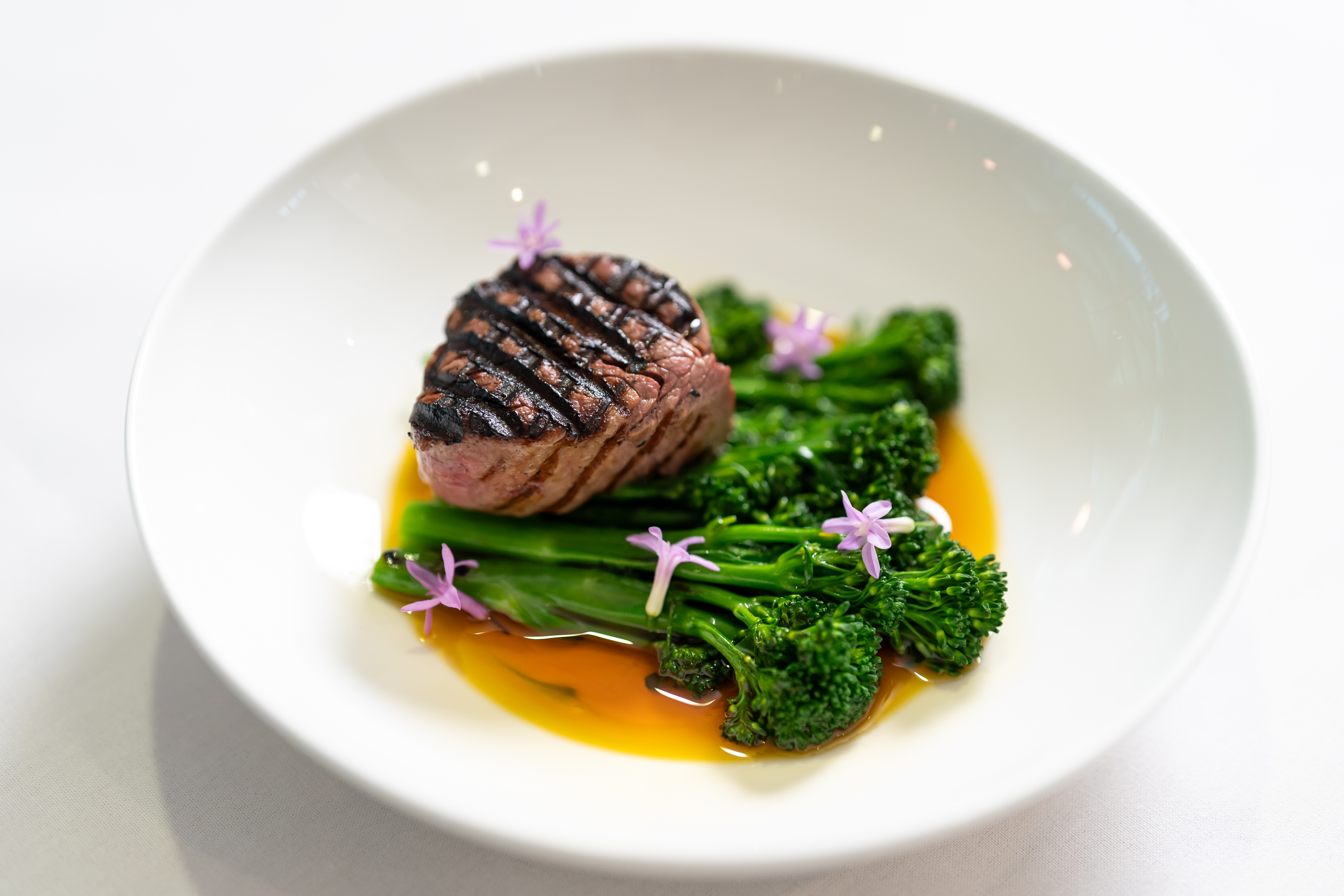 A plated dish featuring a grilled piece of meat served on broccolini with edible flowers and sauce.