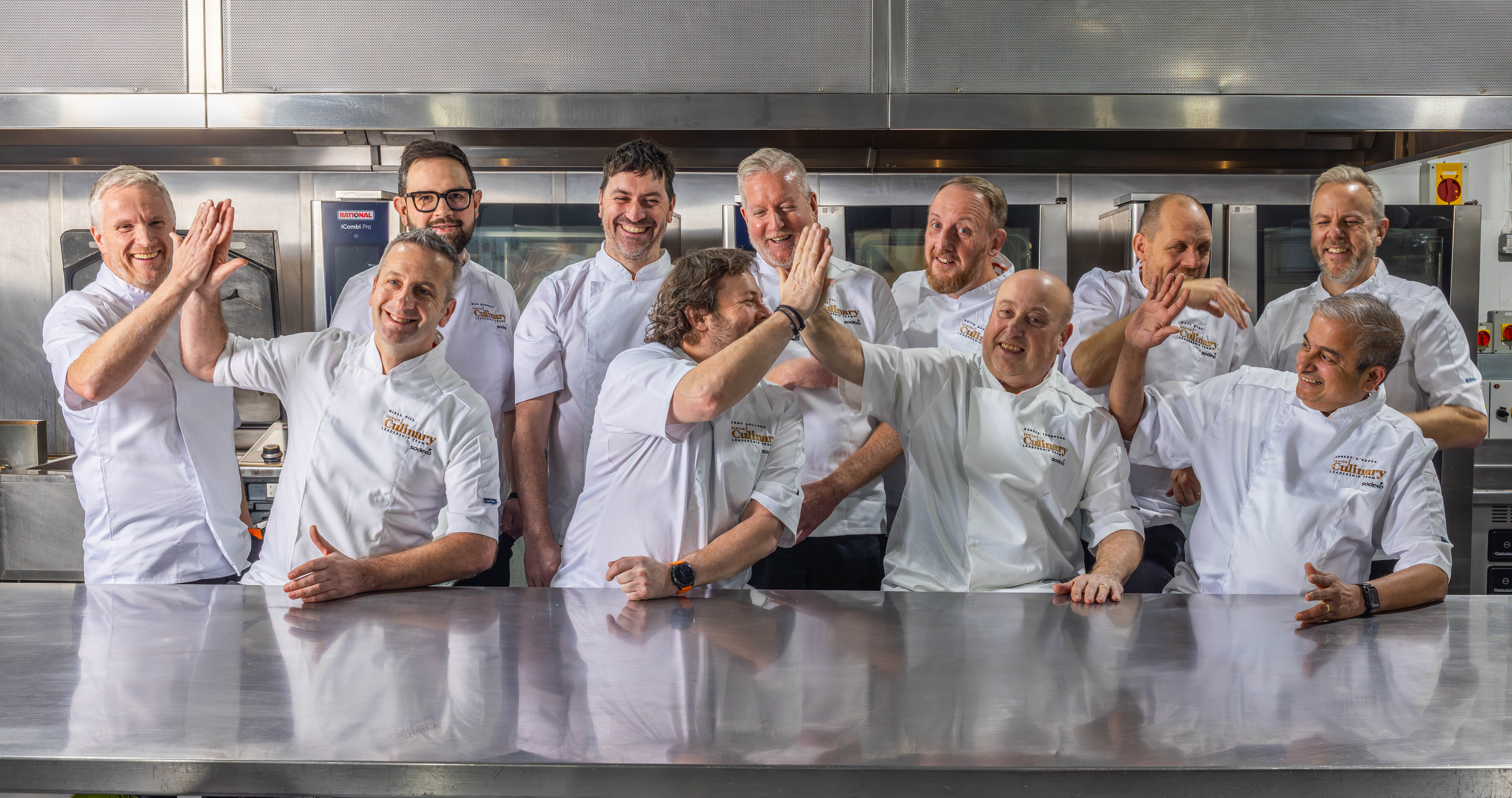 A group of chefs in white uniforms standing together in a commercial kitchen, raising their hands in a lively moment.