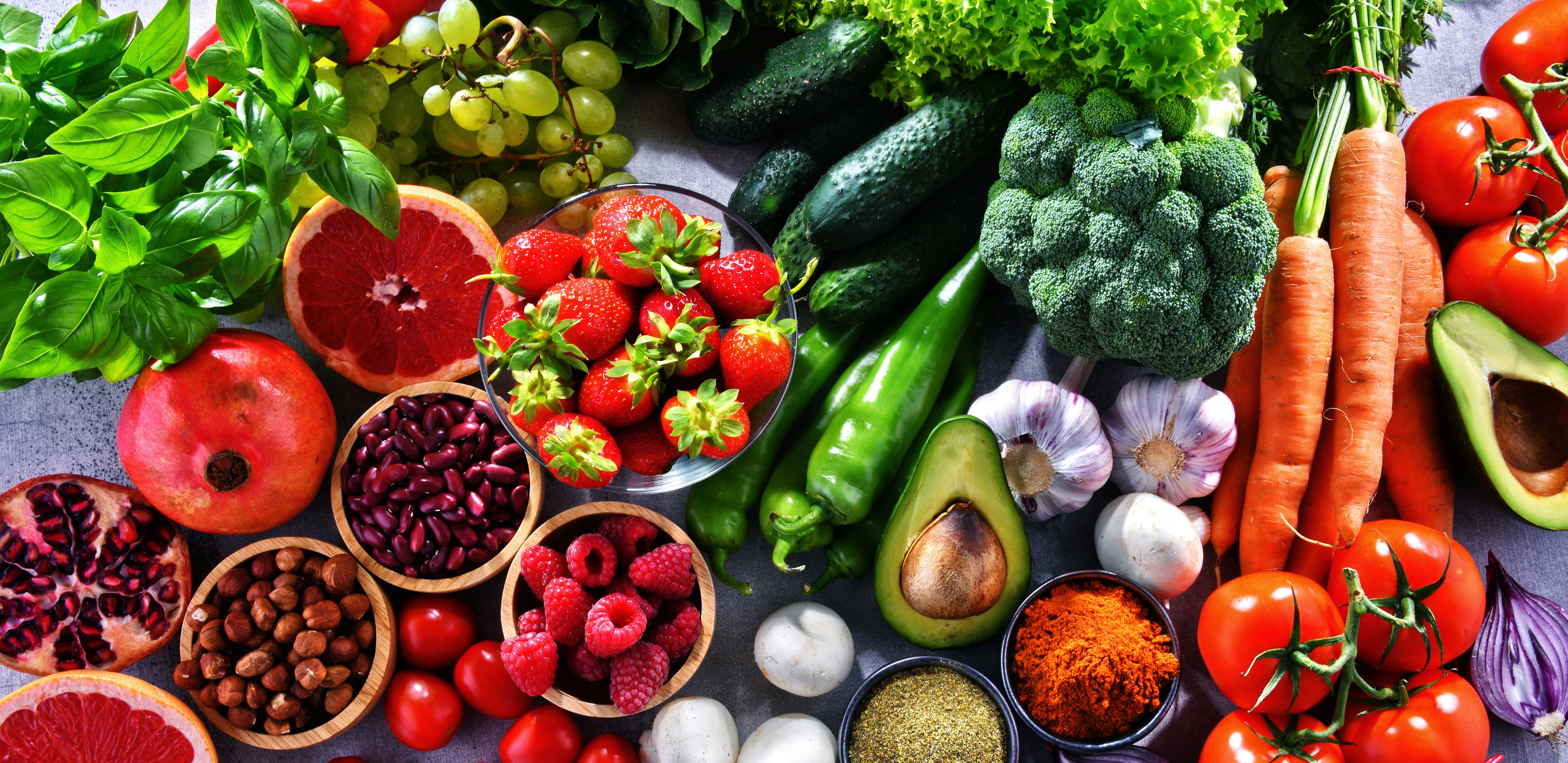Overhead view of a colorful assortment of fresh foods arranged on a gray surface, including tomatoes, carrots, broccoli, cucumbers, green peppers, avocados, garlic, onions, grapes, strawberries, raspberries, pomegranate, grapefruit, basil, nuts, beans, mushrooms, and small bowls of spices