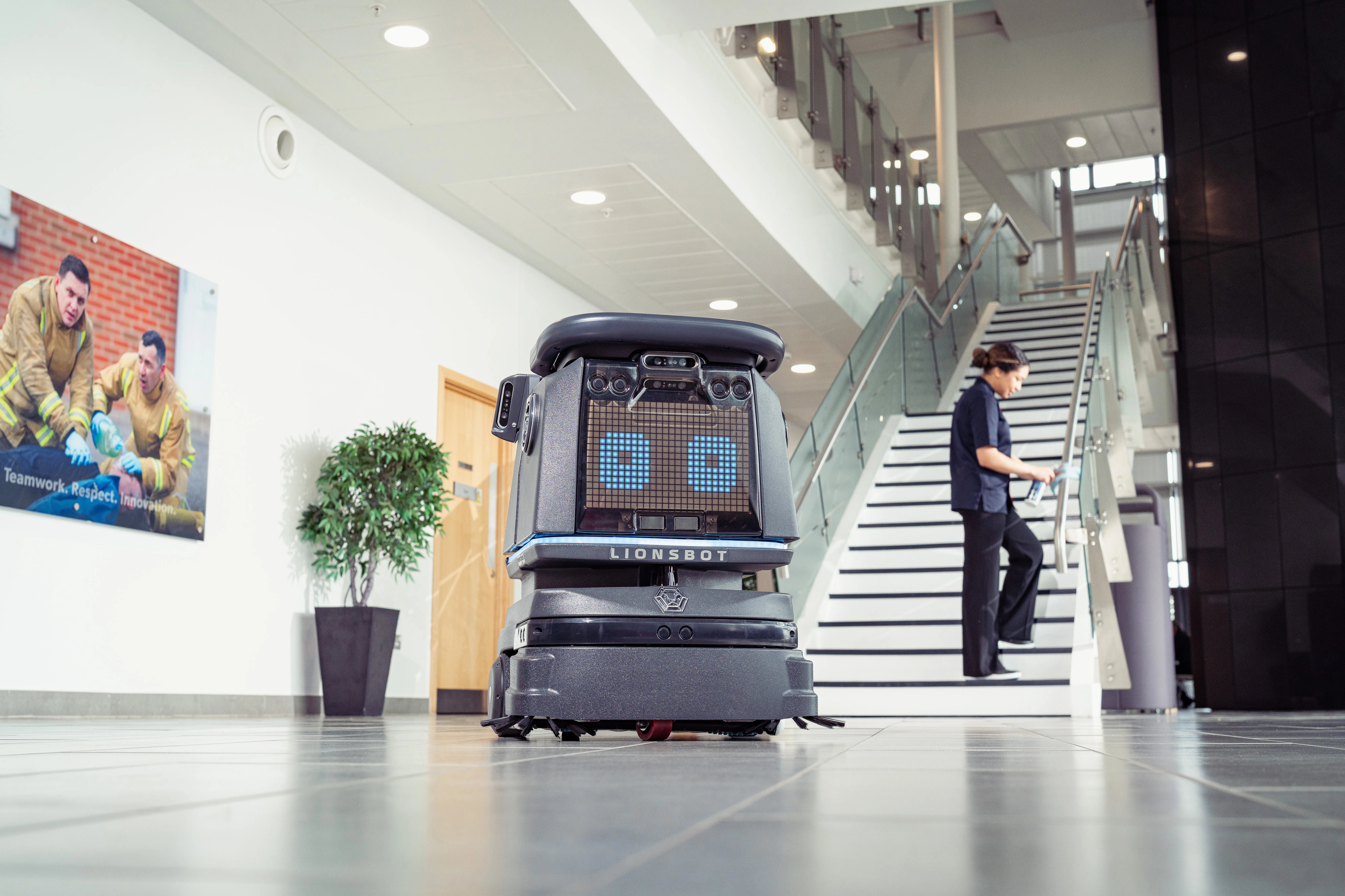 Autonomous cleaning robot operating in a modern office building lobby while a facilities team member works nearby.