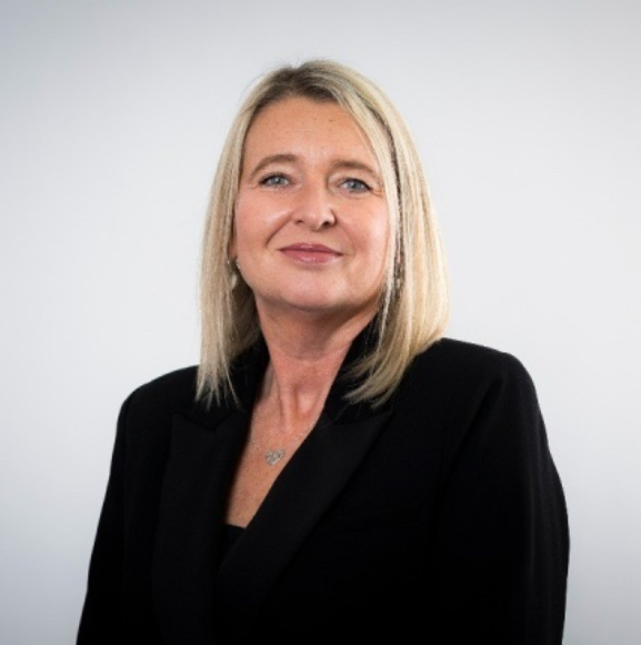 Portrait of a person with shoulder-length light hair wearing a black blazer, standing against a plain light background.