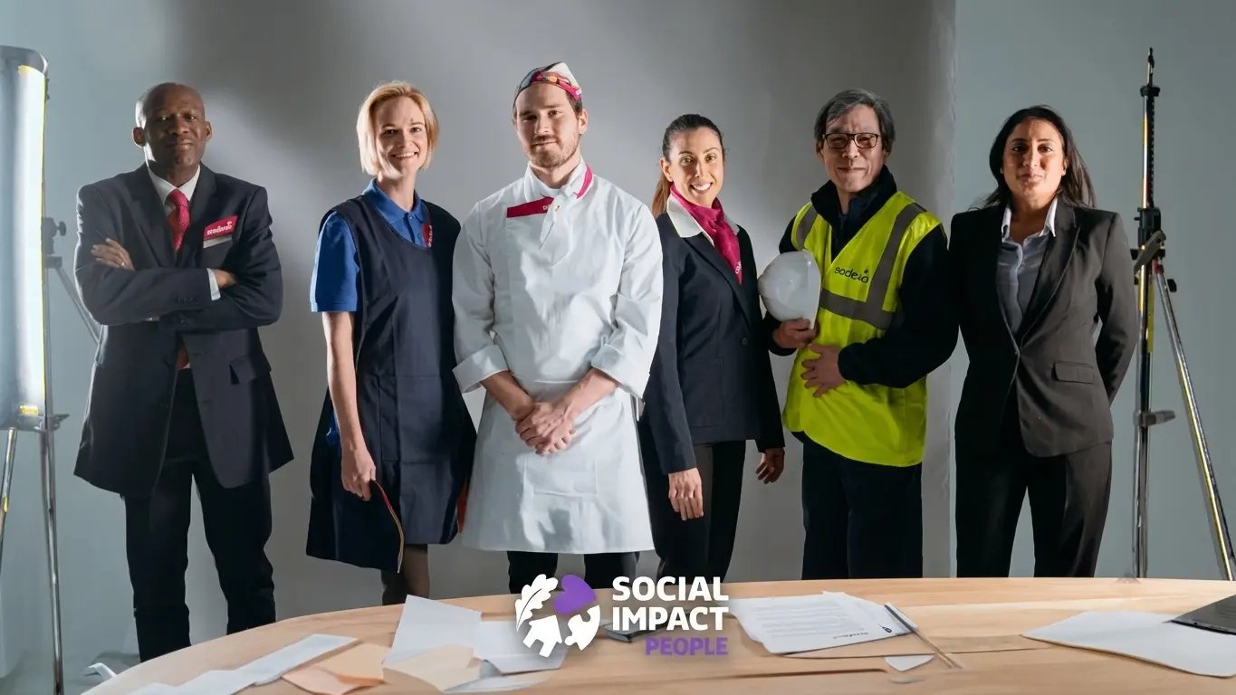 Group of Sodexo team members in different roles, including catering, facilities, and office staff, standing together behind a table in a studio setting with ‘Social Impact People’ branding visible.
