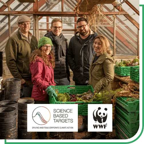 A group of five people standing inside a greenhouse filled with gardening supplies and fresh produce. The group is gathered around green plastic crates containing vegetables such as leafy greens and root vegetables. Shelves and stacks of black plant pots line the left side, while more crates and plants are visible on the right. Sunlight filters through the glass roof and wooden framing of the greenhouse. At the bottom of the image are two logos: one reads “Science Based Targets – Driving Ambitious Corporate Climate Action,” and the other is the WWF panda logo