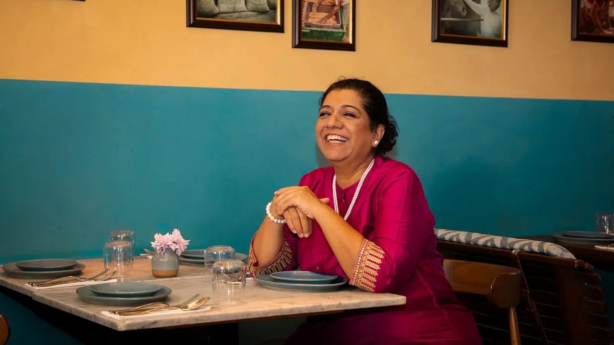 Stop Hunger ambassador Amsa Khan seated at a restaurant table with plates and glasses, wearing a bright pink outfit in a warmly lit dining setting