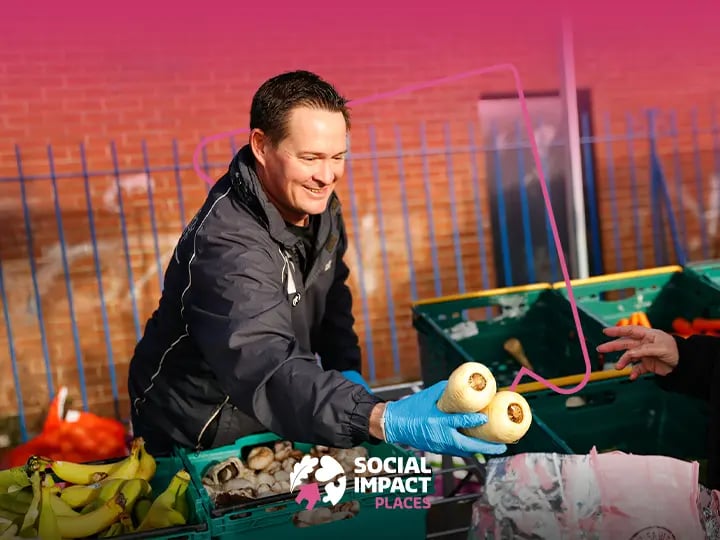 Person wearing blue gloves handing fresh vegetables from green crates at an outdoor food distribution table, with brick wall and railings in the background