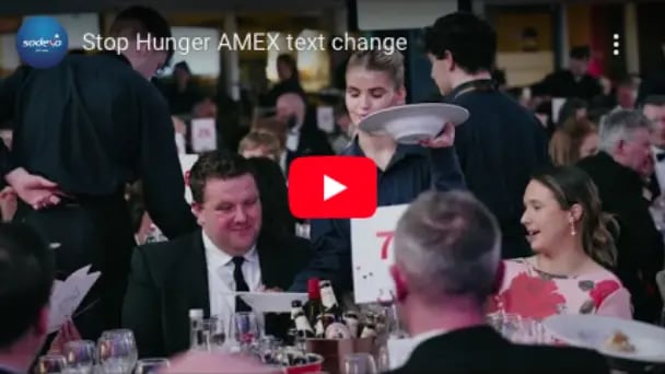Video thumbnail of a formal dinner event with guests seated at tables and a server carrying plates, titled ‘Stop Hunger AMEX text change